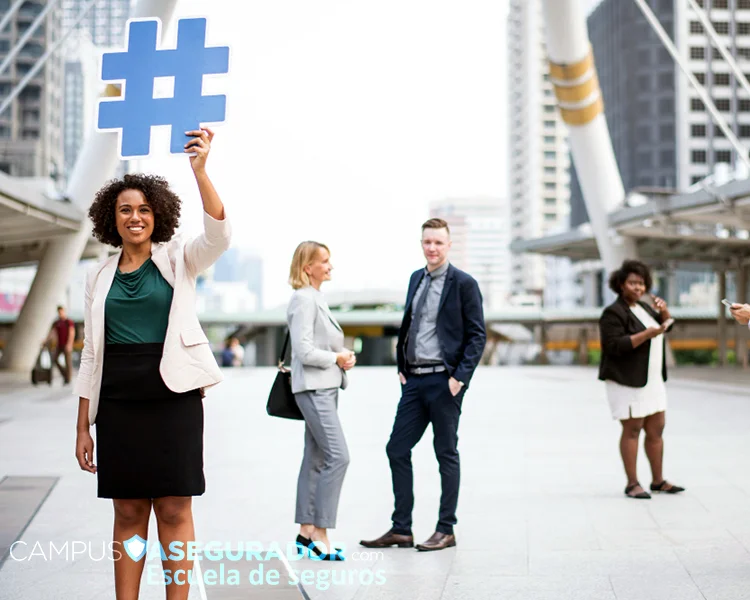 Mujer profesional sostiene hashtag azul de Campus Asegurador en entorno urbano rodeada de ejecutivos de seguros.