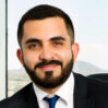 Hombre con barba y traje oscuro sonríe frente a ventanal con fondo de montañas difusas. Joachim-Fayed.jpg
