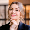 Retrato de mujer profesional con blazer oscuro y mano en mentón sobre fondo desenfocado de interiores.