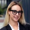 Mujer profesional con cabello rubio, blazer negro y gafas estilo cat-eye sonriendo en un entorno urbano desenfocado.