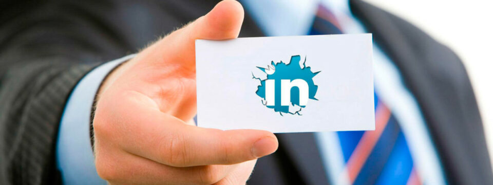 Mano en traje sostiene tarjeta blanca con logotipo de LinkedIn emergiendo mediante un efecto de ruptura central azul.