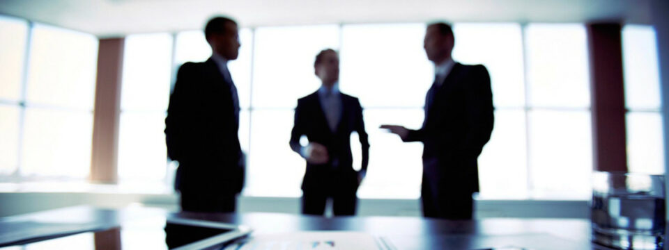 Tres ejecutivos en traje silueteados conversando en una oficina luminosa frente a ventanales en reunión de negocios.