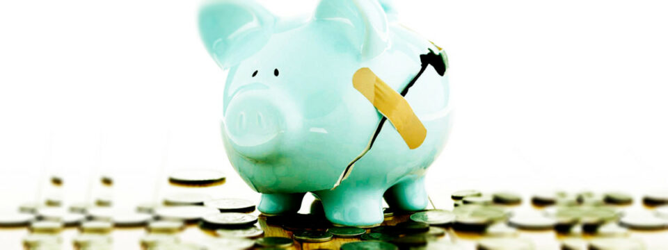 Hucha de cerdo azul agrietada con un apósito adhesivo rodeada de monedas sobre fondo blanco para ahorro y finanzas.