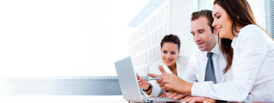 Tres profesionales, dos mujeres y un hombre, colaboran frente a una laptop en oficina corporativa moderna e iluminada.