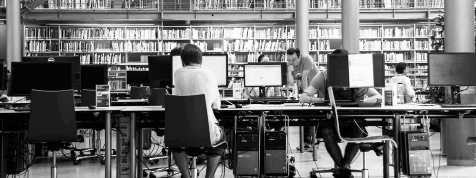 Investigadores en puestos informáticos de una biblioteca moderna con amplias estanterías de libros y diseño industrial.