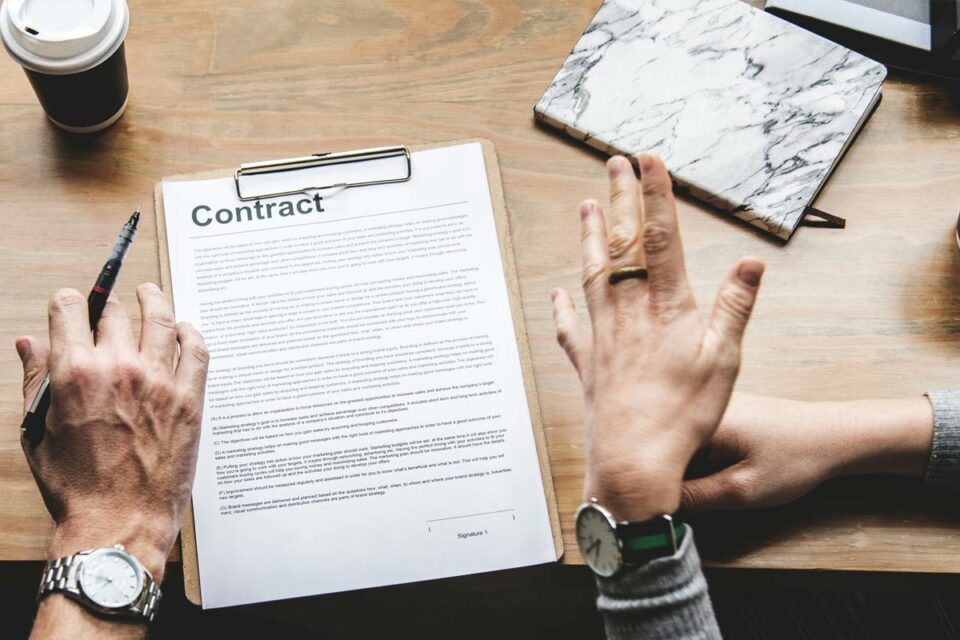 Manos negociando sobre contrato legal impreso en escritorio madera con bolígrafo, reloj pulsera y cuaderno marmolado.