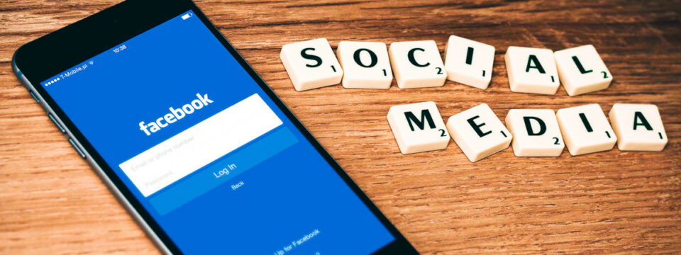 Smartphone mostrando login de Facebook junto a letras de madera que deletrean SOCIAL MEDIA sobre superficie rústica.
