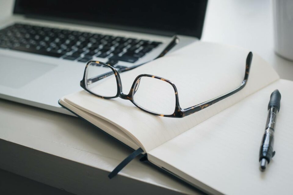 Gafas con montura carey sobre cuaderno abierto junto a laptop y bolígrafo en escritorio de oficina minimalista.