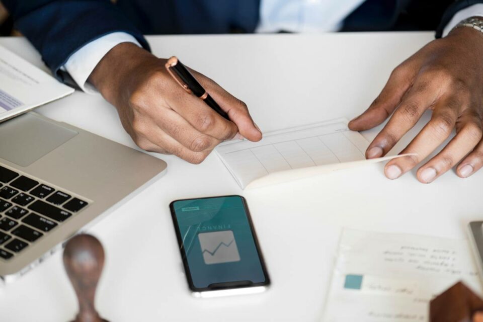 Persona de negocios firmando documento bancario junto a smartphone con aplicación de finanzas y gráfico de inversión.