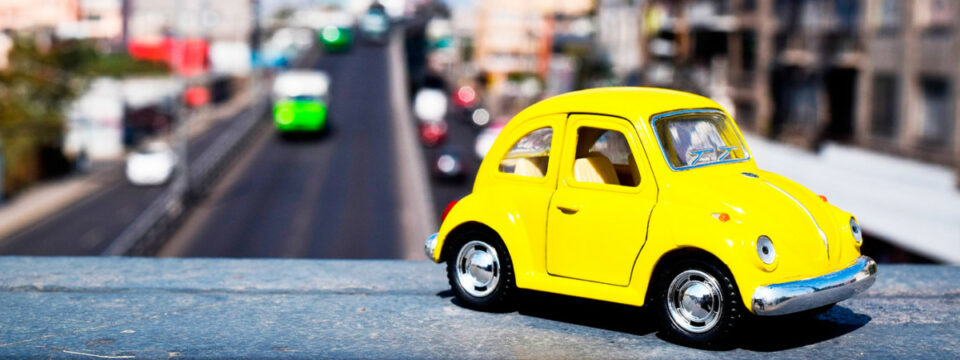Coche miniatura Volkswagen Beetle amarillo posado en repisa frente a tráfico urbano con fondo desenfocado.