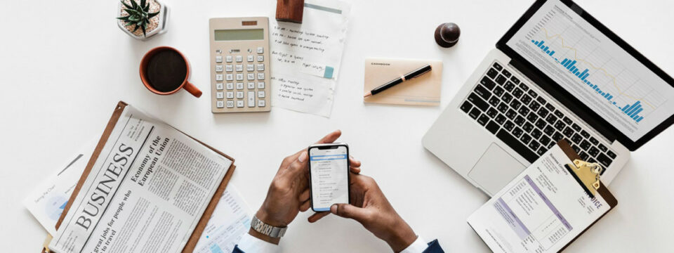 Vista cenital de manos usando smartphone sobre escritorio con laptop, gráficos financieros, calculadora y prensa económica.