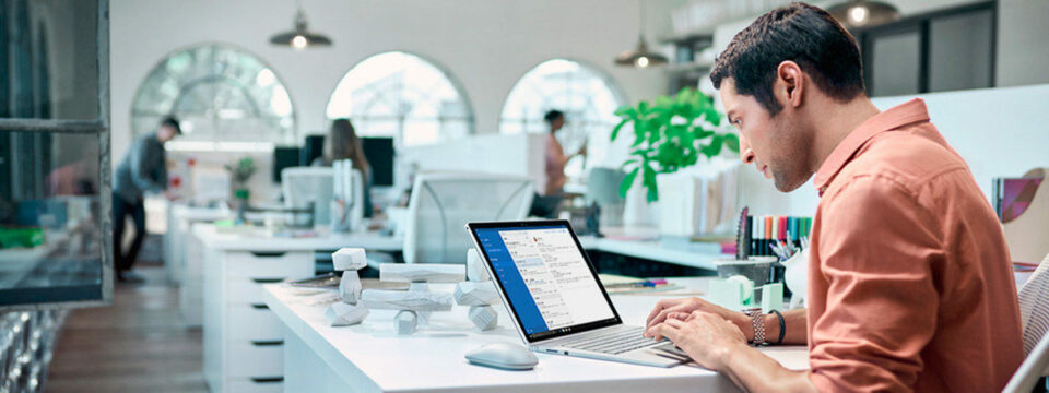 Profesional trabajando en laptop Microsoft Surface dentro de oficina moderna con iluminación natural y diseño minimalista.