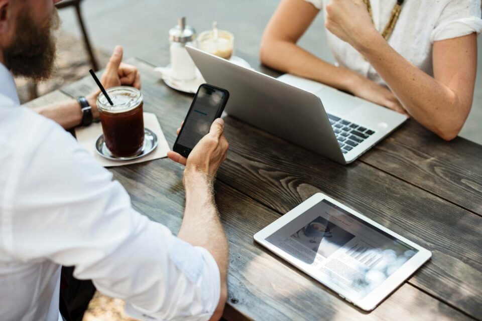 Hombre usando smartphone y mujer con laptop en mesa de madera exterior con tablet y café frío en entorno de coworking.