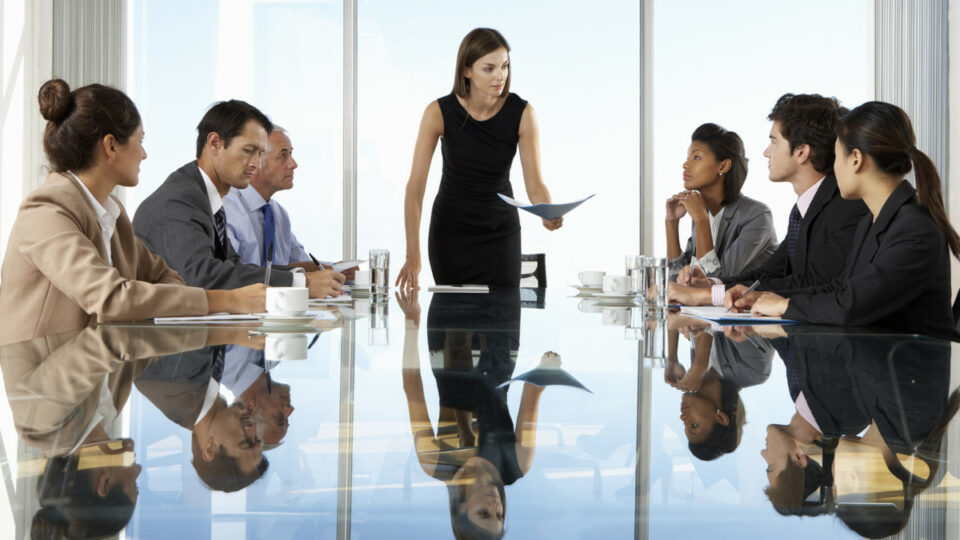 Ejecutiva liderando reunión estratégica con equipo profesional en sala de juntas moderna con superficie reflectante.