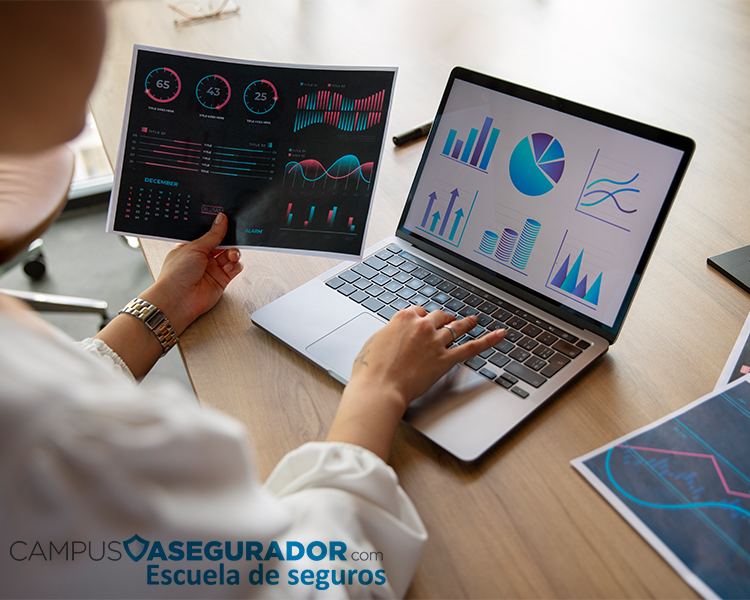 Profesional analizando gráficos de datos en laptop y reportes impresos para formación en Escuela de Seguros Campus Asegurador.