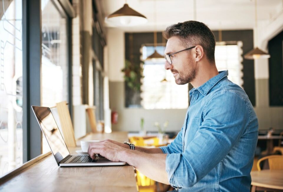 Hombre con gafas y camisa denim operando una laptop en la barra de madera de una cafetería iluminada.