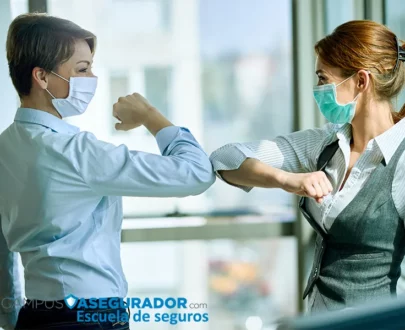 Dos mujeres profesionales con mascarillas quirúrgicas realizando un saludo de codo en entorno de oficina corporativa.