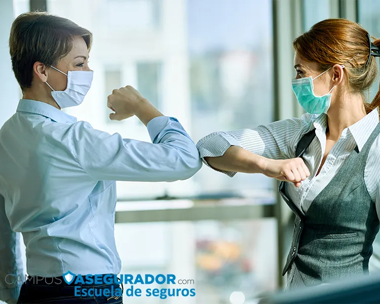 Dos mujeres profesionales con mascarillas quirúrgicas realizando un saludo de codo en entorno de oficina corporativa.