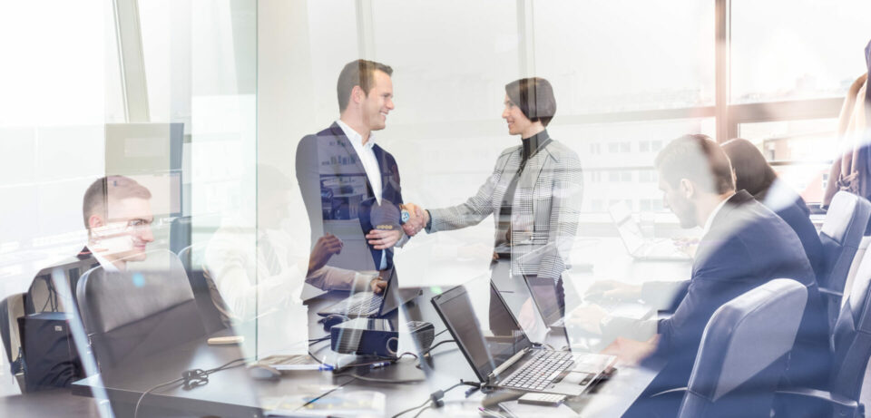 Profesionales en oficina moderna estrechando manos durante reunión de negocios con equipo trabajando de fondo.