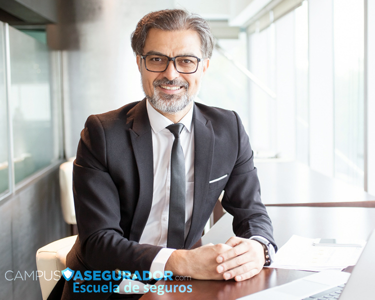 Profesional en traje y gafas sentado en oficina corporativa con logotipo de Campus Asegurador Escuela de Seguros.