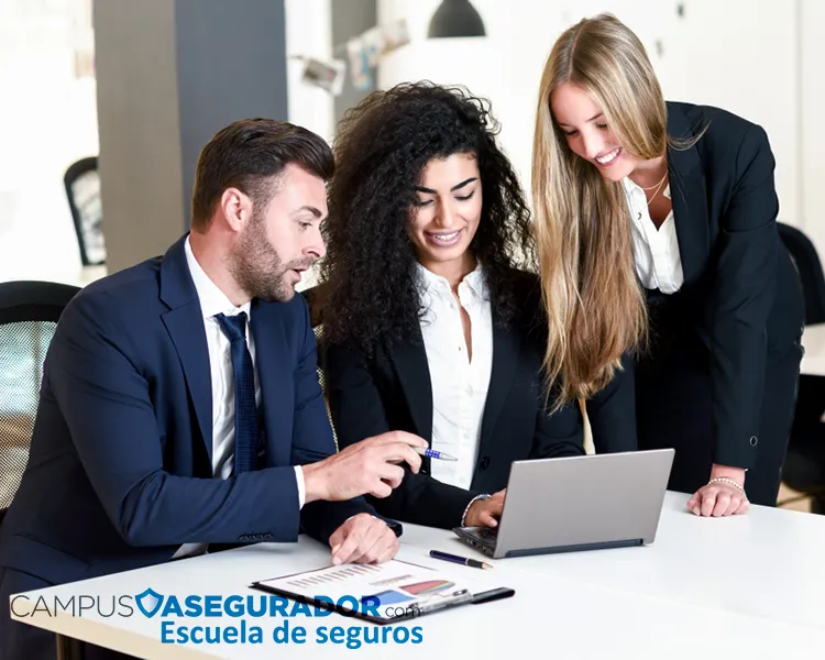 Tres profesionales colaborando con laptop y gráficos impresos en Campus Asegurador, escuela técnica de seguros.
