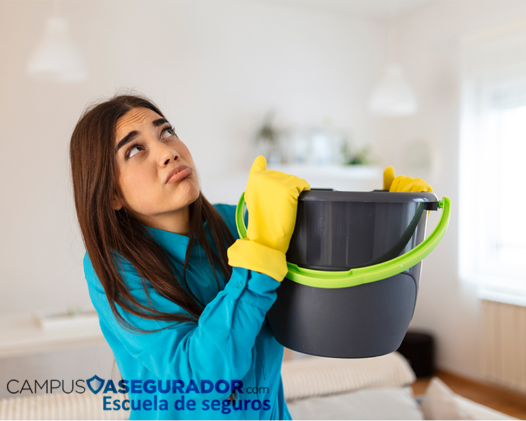 Mujer preocupada con cubo y guantes ante gotera en techo, representando siniestro por daños de agua en vivienda.