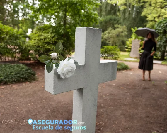 Cruz de piedra con rosas blancas en cementerio, mujer de luto con paraguas al fondo. Logo Campus Asegurador.