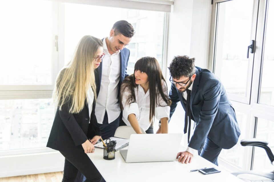 Cuatro profesionales analizan datos en una laptop en una oficina corporativa con iluminación natural y ventanales amplios.