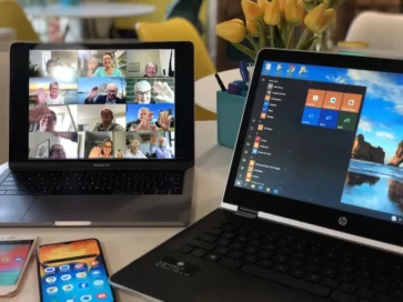 Adultos mayores en videoconferencia grupal desde MacBook Pro, con laptop HP y smartphones sobre escritorio con flores.