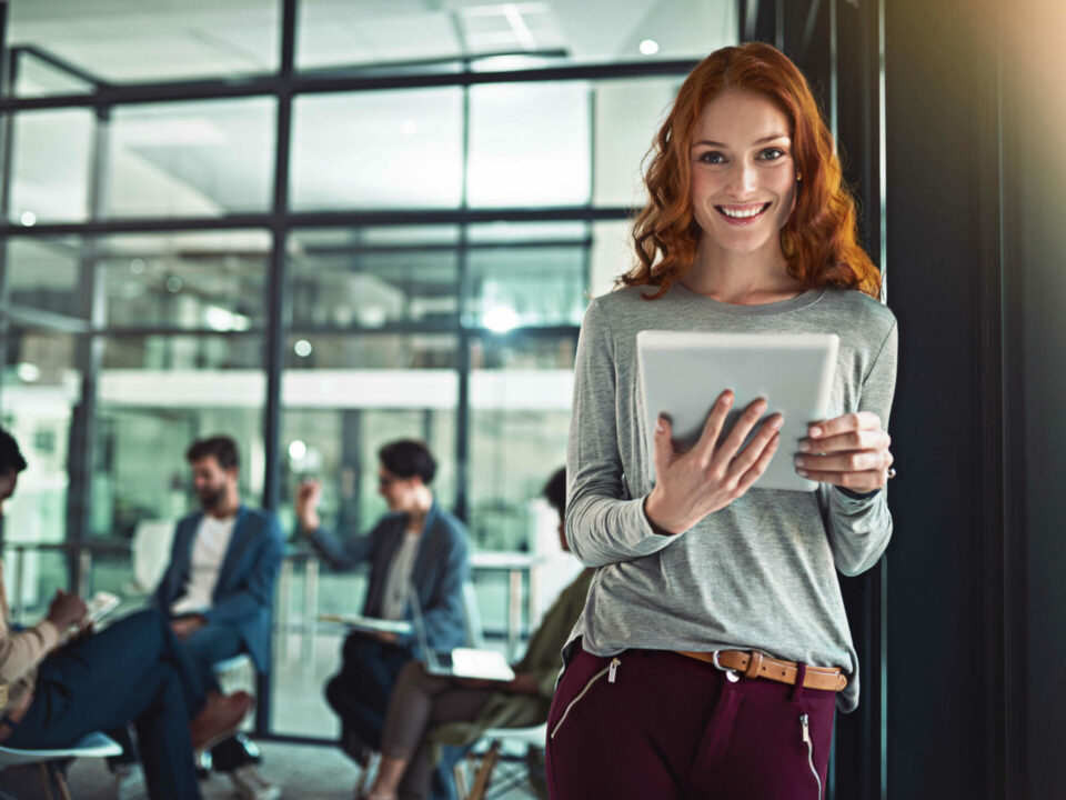 Mujer pelirroja sonriente operando tablet digital en oficina corporativa moderna con equipo de trabajo al fondo.