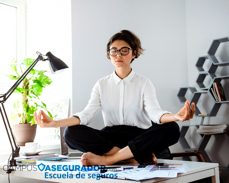 Mujer profesional meditando en posición de loto sobre escritorio de oficina junto al logo Campus Asegurador.