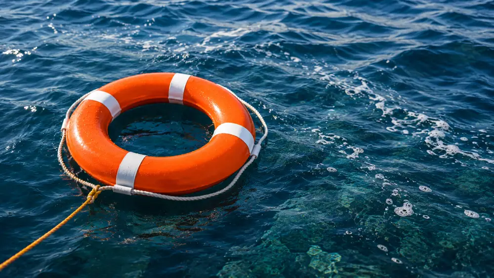 Aro salvavidas naranja con bandas blancas reflectantes sobre superficie de agua marina ondulada con cabo de amarre.