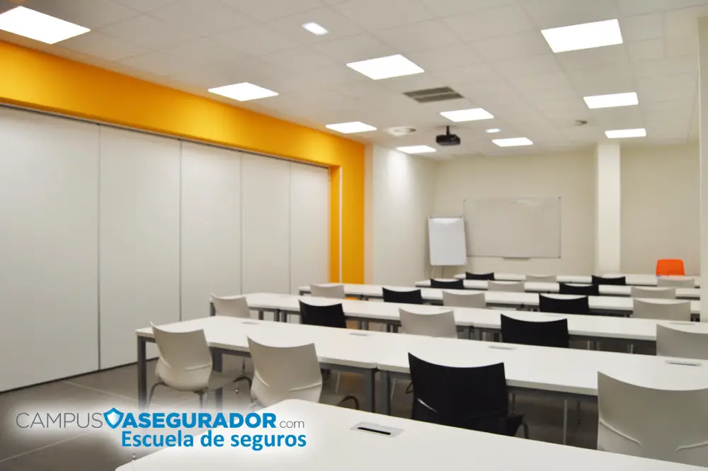 Aula de Formación Seguros – Campus Asegurador Aula técnica Campus Asegurador con mobiliario ergonómico blanco y negro, proyector y panel naranja corporativo.