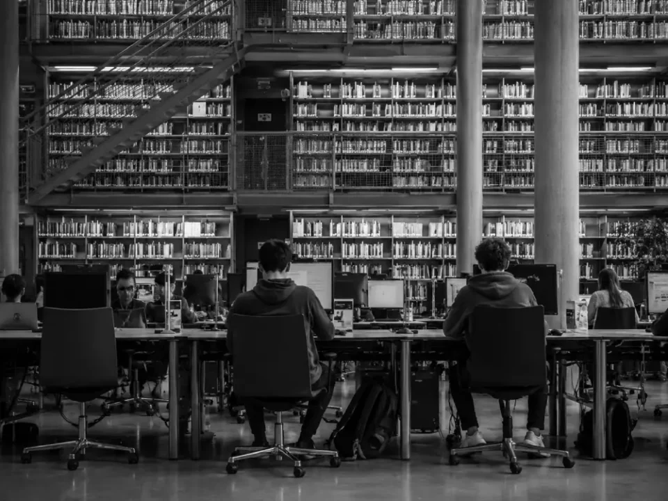 Investigadores en puestos informáticos de una biblioteca moderna con amplias estanterías de libros y diseño industrial.