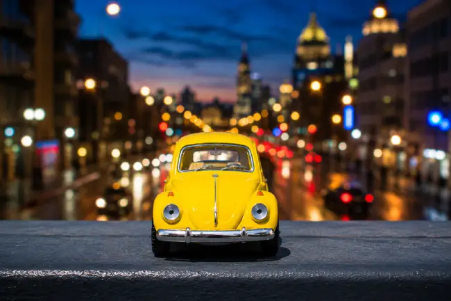 Miniatura Volkswagen Beetle Amarillo: Fotografía Urbana Coche miniatura Volkswagen Beetle amarillo posado en repisa frente a tráfico urbano con fondo desenfocado.