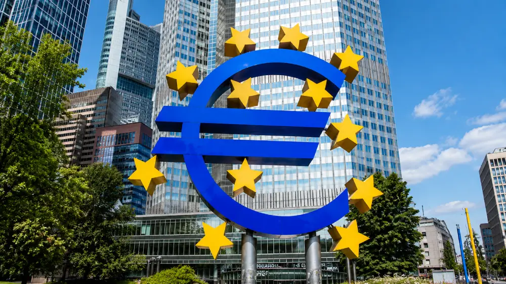 Escultura azul del símbolo euro rodeada de estrellas amarillas frente a rascacielos en Frankfurt.