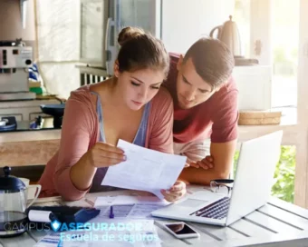 Pareja joven revisa documentos financieros junto a portátil y calculadora en mesa de cocina con logo Campus Asegurador.